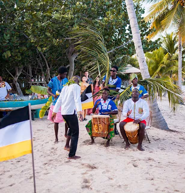 Garifuna drummers and dancers performing on the beach beside a Garifuna flag, with guests watching